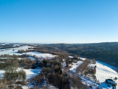 Açık mavi gökyüzünün altındaki karla kaplı tarlaların ve ağaçların havadan görünüşü sakin bir kış manzarası sergiliyor. Doğayla ilgili projeler için mükemmel.