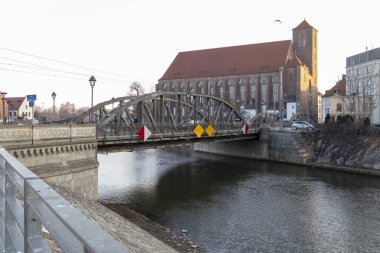 Wroclaw Polonya - 23 Şubat 2025: Eski bir tuğla kilise ve kentsel manzaranın yanı sıra görünür uyarı levhalarıyla birlikte tarihi bir metal köprü nehir boyunca uzanıyor