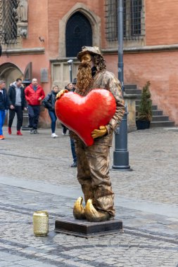Wroclaw Polonya - 23 Şubat 2025: Bronz elbiseli bir sokak sanatçısı, canlı bir şehir meydanında yoldan geçenleri büyüleyen büyük kırmızı bir kalp tutuyor.