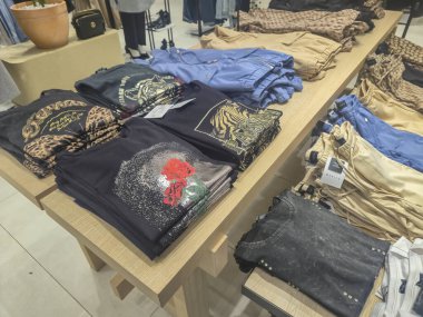 Przemysl Poland - Mart 12025: Grafik tişörtler ve perakende satış mağazasındaki ahşap masalarda düzgünce düzenlenmiş günlük pantolonlar da dahil olmak üzere çeşitli giyim eşyalarının gösterimi.