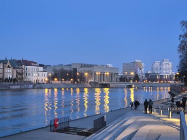 Wroclaw Polonya - 23 Şubat 2025: Aydınlatılmış binalar ve mavi gökyüzü altında gezinen yayaların olduğu Odra Nehri rıhtımının akşam manzarası.