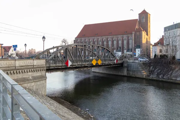 Wroclaw Polonya - 23 Şubat 2025: Eski bir tuğla kilise ve kentsel manzaranın yanı sıra görünür uyarı levhalarıyla birlikte tarihi bir metal köprü nehir boyunca uzanıyor