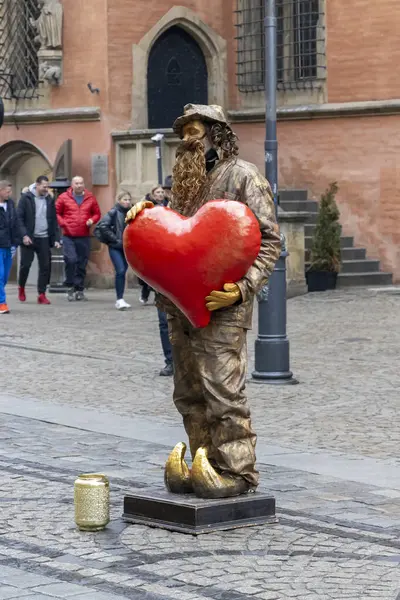 Wroclaw Polonya - 23 Şubat 2025: Bronz elbiseli bir sokak sanatçısı, canlı bir şehir meydanında yoldan geçenleri büyüleyen büyük kırmızı bir kalp tutuyor.