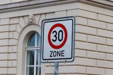 30 bölge hız limitini gösteren bir trafik levhası klasik bir binanın önünde duruyor. Çevre, şehrin işlek bir sokak atmosferine işaret ediyor.
