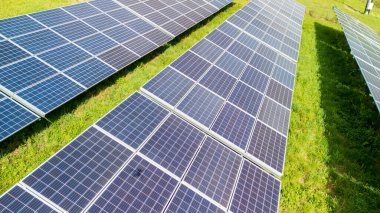 Güneş panellerinin hava görüntüsü sıralanarak yenilenebilir enerji üretimi için güneş ışığını yakalıyor. Açık mavi gökyüzünün altında sürdürülebilirliği destekleyen çimenli bir arazide yer alıyor..
