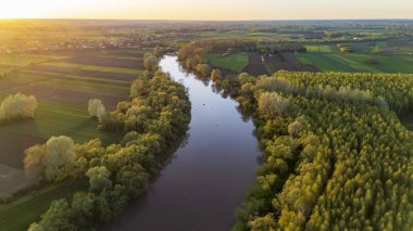 Gün batımında verimli yeşillikler arasında süzülen sakin bir nehrin havadan görünüşü. Etrafı tarım arazileri ve ağaçlarla çevrili. Güneşin sıcak parıltısı manzaraya huzurlu bir atmosfer ekler..