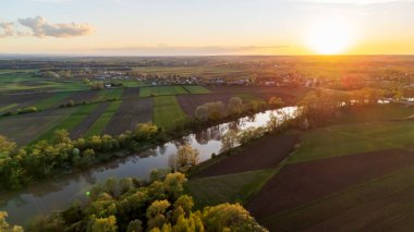 Gün batımında sakin bir manzaranın havadan görünüşü. Bereketli tarlalar, dolambaçlı bir nehir ve uzak bir köy. Bahar mevsiminde çekilen canlı renkler çerçeveye hakim olur.