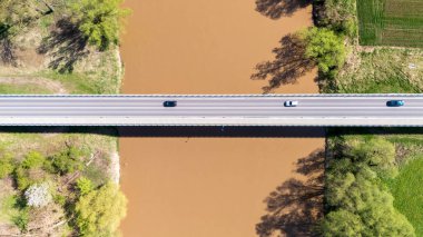 Çamurlu bir nehri kaplayan ve araçlarla seyahat eden bir köprünün havadan görüntüsü. Yeşillik, doğa ve altyapıyı betimleyen nehir kıyılarını çevrelemektedir..