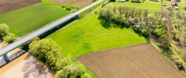 Bereketli yeşil tarlalar ve bir köprünün altındaki kıvrımlı bir nehir baharda kırsal alanların güzelliğini gösteriyor ve tarımsal gelişimi vurguluyor..