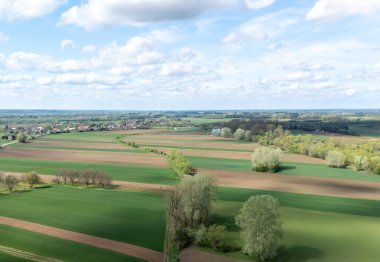 Parçalı bulutlu bir gökyüzünün altındaki tarım arazileri ile karışmış yemyeşil tarlaların havadan görünüşü. Manzara ilkbaharda kırsal huzuru sergiliyor.