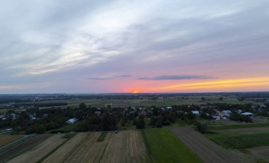 Gün batımında sakin bir manzaranın havadan görünüşü tarlalar ve sıcak turuncu ve mor renklerde yıkanmış küçük bir köy huzurlu bir kırsal manzarayı yansıtıyor..