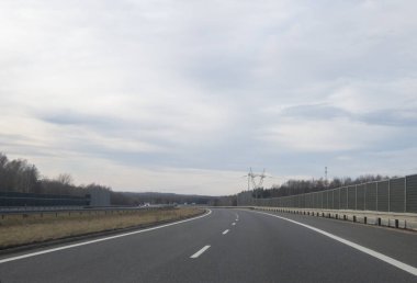 Açık otoyol, bulutlu bir gökyüzünün altında ses bariyerleri ve elektrik hatlarıyla çevrili mesafeye kadar uzanıyor. Bu sahne özgürlük ve seyahat hissi uyandırıyor..