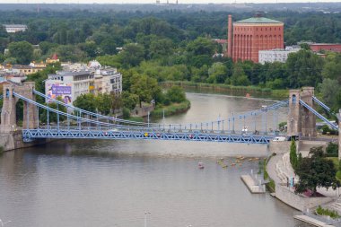 Wroclaw Polonya - 16 Haziran 2022: Nehir üzerindeki asma köprünün manzarası yeşil binalar ve kayakçılarla manzaranın keyfini çıkarıyor.