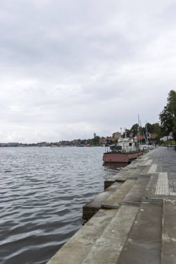 Mikolajki Polonya - 29 Eylül 2024: Bulutlu bir gökyüzünün altında ağaçlarla çevrili teknelerle dolu bir göl manzarası.