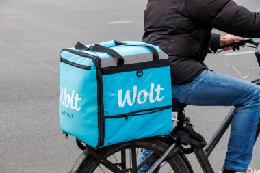 Berlin Almanya - 11 Nisan 2025: Bir bisikletçi, sokak ortamında bisiklet sürerken parlak mavi bir Wolt teslimat çantası kullanarak yiyecek dağıtıyor.