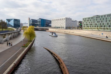 Berlin Almanya - 11 Nisan 2025: Nehir kıyısı boyunca modern binaların sergilendiği şehir manzarası, bulutlu bir günde yürüyen insanlar ve denizde yüzen bir tekne.