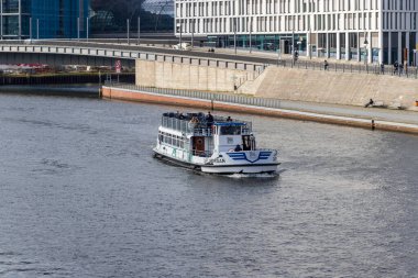 Berlin Almanya - 11 Nisan 2025: Spree Nehri boyunca modern mimariye yakın seyreden yolcularla birlikte bir nehir teknesi şehir manzarası sunuyor.