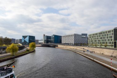 Berlin Almanya - 11 Nisan 2025: Canlı yeşil ağaçlarla dolu bir nehir kıyısında modern binaların manzarası. Bir köprü her iki tarafı da gezinti güvertesinde yürüyen insanlarla birleştirir..
