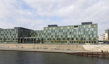 Berlin Almanya - 11 Nisan 2025: Deniz kenarındaki yeşil cam binanın modern mimarisi bulutlu gökyüzü dramatik bir zemin ve kentsel atmosfer yaratıyor.