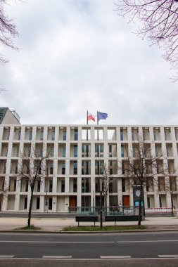 Berlin Almanya - 11 Nisan 2025: Çağdaş mimariyi gösteren bulutlu bir atmosferde ağaçlarla çevrili Polonya ve AB bayraklarının yer aldığı modern bir bina.