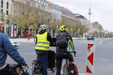Berlin Almanya - 11 Nisan 2025: Şehir caddesinde trafik ışıklarında bekleyen iki bisikletçi. Dükkanları boş yollar ve ilkbahar ağaçları olan şehir manzarası.