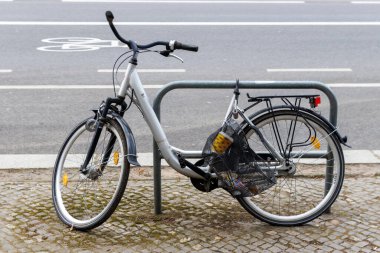 Berlin Almanya - 11 Nisan 2025: Bisiklet parkında güvenli bir şekilde park edilmiş gümüş bir bisiklet bisiklet yolu ve yaya kaldırımının arka planına karşıdır.