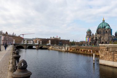 Berlin Almanya - 11 Nisan 2025: Spree Nehri boyunca uzanan tarihi Berlin Katedrali 'nin bulutlu bir günde yürüyen yayalarla manzarası.