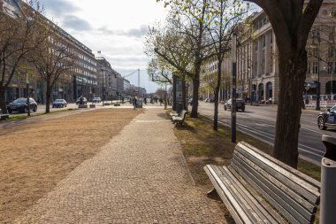 Berlin Almanya - 11 Nisan 2025: Bulutlu bir gökyüzü altında ağaçlar ve binalarla dolu bir patika boyunca uzanan sakin bir şehir sokak sahnesi.