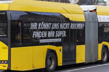 Berlin Almanya - 11 Nisan 2025: Sarı elektrikli bir otobüs, şehir sokaklarında araba sürerken çevre dostu özelliklerini tanıtan neşeli bir slogan içeren bir reklam sergiliyor.