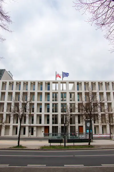 Berlin Almanya - 11 Nisan 2025: Çağdaş mimariyi gösteren bulutlu bir atmosferde ağaçlarla çevrili Polonya ve AB bayraklarının yer aldığı modern bir bina.