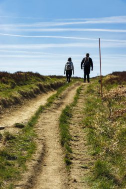 İki yürüyüşçü, açık mavi gökyüzünün altındaki dağlarda manzaralı bir toprak yolda yürüyor. Doğada güneşli bir günün tadını çıkarıyor. Açık hava aktivitelerini ve macerayı teşvik ediyorlar..