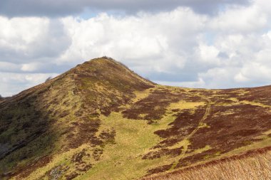 Bulutlu bir gökyüzünün altındaki yuvarlanan tepelerin manzarası yemyeşil ve kahverengi yamalar sergiliyor. Açık hava keşfi ve eğlence için mükemmel sakin bir manzara..