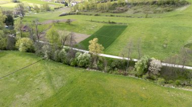 İlkbaharın başlarında çayırların ve ağaçların bulunduğu yemyeşil bir arazinin havadan görünüşü. Sahne canlı renkleri, açık alanları ve sakin kırsal atmosferi yakalıyor..