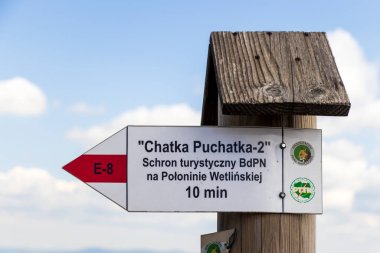 Polonya 'nın Bieszczady Dağları' ndaki Chatka Puchatka barakasına giden yolu gösteren işaret direği. İşaret, parlak bir gökyüzüne karşı belirlenmiş uzaklık bilgisi ve doğa sembollerini içeriyor..