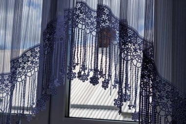 Dekoratif dantel perdeler zarif bir şekilde pencerenin üzerine örtülmüş. Karmaşık gölgeler bırakıyor. Doğal ışık, kumaş ve açık hava manzarası arasında canlı bir kontrastı ortaya çıkarır..