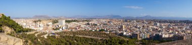 Alicante İspanya 'nın panoramik manzarası berrak mavi gökyüzü altında tepeler ve dağlarla çevrili yerleşim ve ticari binalarla dolu canlı bir şehir manzarası sergiliyor..