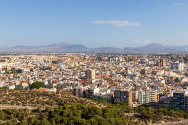 Alicante İspanya 'nın panoramik manzarası çeşitli şehir mimarisi yeşil tepeler ve uzak dağlar gün ışığında açık mavi bir gökyüzü altında.