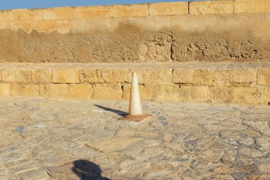 Açık havada, açık mavi bir gökyüzünün altında yıpranmış bir duvara karşı taş bir kaldırıma koni konulmuş. Olay yerinde inşaat veya güvenlik önlemleri alınıyor.