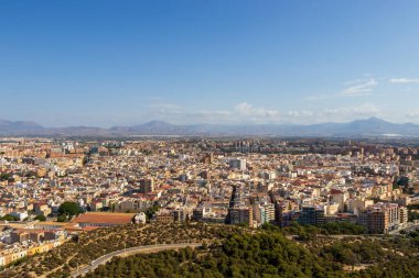 Alicante İspanya 'nın hava görüntüsü açık mavi gökyüzünün altında yeşil binalar ve dağlarla dolu canlı bir şehir manzarası sergiliyor. Seyahat ya da şehir hayatı temaları için mükemmel.