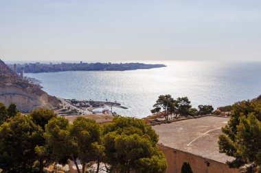 Berrak bir gökyüzü altında mavi sular ve kentsel ufuk çizgisinin yer aldığı Alicante İspanya kıyılarının panoramik bir görüntüsü. Seyahat ve turizm temaları için ideal.
