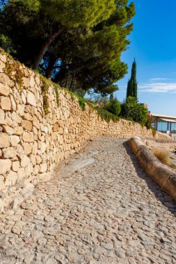 Sade bir kaldırım taşı yolu taştan bir duvar ve yeşil alanla kaplı. Ziyaretçileri berrak mavi gökyüzünün altında, huzurlu bir açık hava ortamından geçiriyorlar..