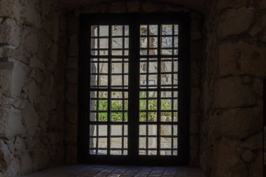 Koyu bir demir ızgara penceresinin içindeki taş bir duvarın çerçevelenmiş görüntüsü. Dışarıdan görünen yeşillik ipuçları. Huzurlu bir ortamda karanlıkla aydınlık arasında bir tezat oluşturuyor..