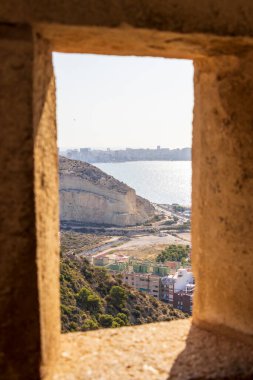 Sahil şeridine ve Alicante plajına bakan tarihi bir taş pencereden bakıldığında güneşli bir günde sakin mavi denizi ve modern binaları yakalıyor..