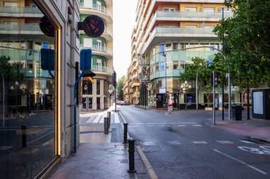 Alicante İspanya - 11 Temmuz 2025: Alicante 'de sakin bir kent sokak sahnesinde kaldırımlarda yayalar ve ağaçların oluşturduğu modern mimari sergileniyor.