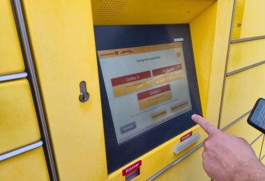 Lahde Almanya - 15 Nisan 2025: Kullanıcı bir DHL paket dolabının dijital ekranıyla etkileşime geçiyor paket boyutu seçenekleri seçiliyor ve elinde akıllı telefon bulunuyor.