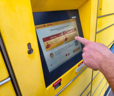 Lahde Almanya - 15 Nisan 2025: Bir kullanıcı şehir ortamlarında modern lojistik teknolojisini gösteren bir paket almak için DHL paket istasyonu dokunmatik ekranı ile etkileşime girer.