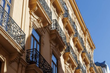 Valencia 'daki zarif bir binanın, parlak mavi gökyüzüne karşı süslü balkonları ve dekoratif unsurları gösteren cephesinin detaylı bir görüntüsü. Mimari projeler için ideal.