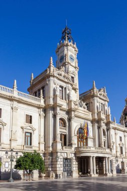 Alicante 'deki belediye binası, açık mavi gökyüzünün altında duran belirgin bir saat kulesinin yerel yönetim ve kültür mirasını temsil ettiği karmaşık bir mimariyi gözler önüne seriyor..