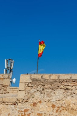 İspanya bayrağı Alicante 'de Castillo de Santa Barbara' da açık mavi gökyüzüne karşı dalgalanıyor. Tarihi eserde taş mimari ve kültür mirası sergileniyor..