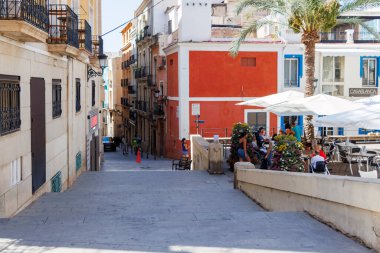 Alicante İspanya - 12 Temmuz 2025: Alicante 'de renkli binaların, kalabalık bir meydanın ve sıcak güneşli bir atmosferde insanların yemek yediği canlı bir sokak manzarası.